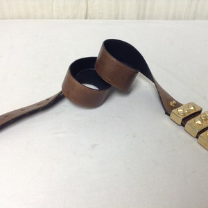 May include: A brown leather belt with a gold metal buckle featuring three rows of pyramid-shaped studs.