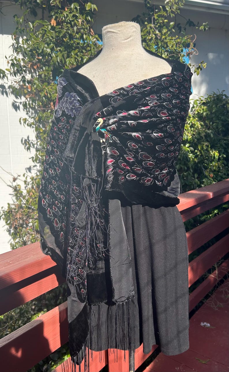May include: Black shawl with a velvet texture and a peacock feather pattern in red and white. The shawl is draped over a black dress form. The shawl has a fringe detail and a peacock brooch.
