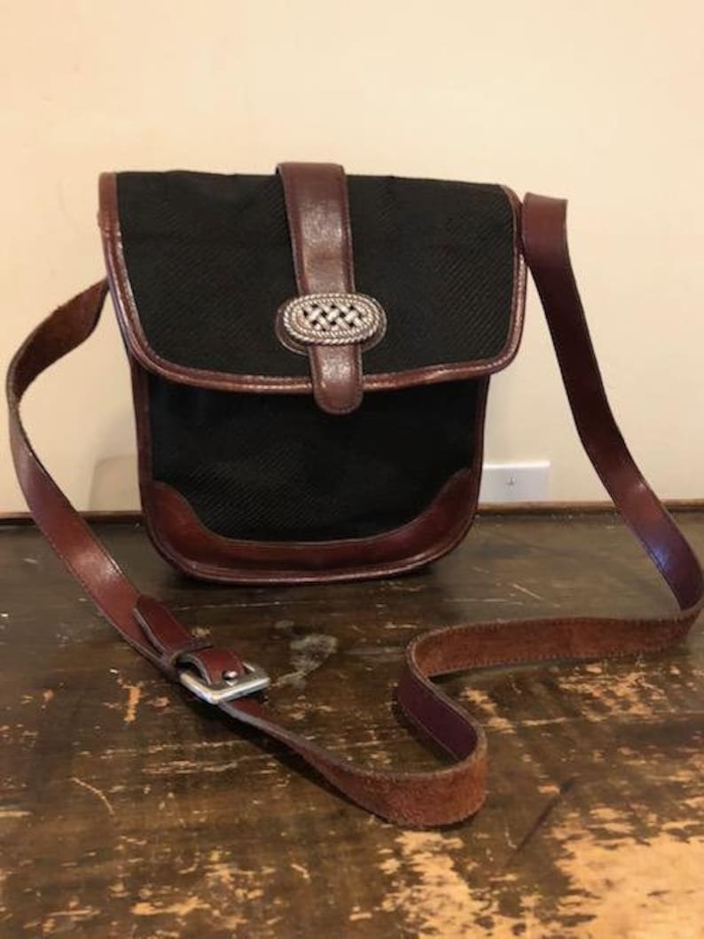 Op de afbeelding: Een zwarte canvas crossbody tas met een bruine leren flap en een zilveren gesp. De tas heeft een lange bruine leren band.