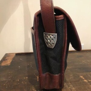 Op de afbeelding: Een zwarte canvas en bruine leren schoudertas met een zilveren metalen sluiting. De tas heeft een lange bruine leren band.