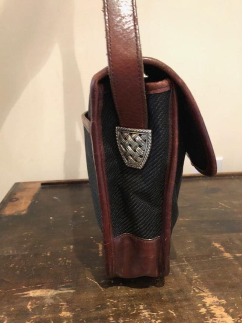 Op de afbeelding: Een zwarte canvas en bruine leren schoudertas met een zilveren metalen sluiting. De tas heeft een lange bruine leren band.