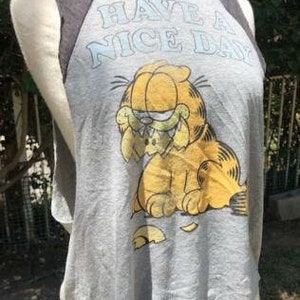 May include: Gray sleeveless t-shirt featuring Garfield the cat with the phrase "HAVE A NICE DAY" in light blue. The shirt has dark gray raglan sleeves and a distressed, vintage look.