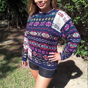 May include: A blue, white, and pink patterned sweater with long sleeves. The sweater has a fair isle design with geometric shapes and a white patch on the right shoulder.
