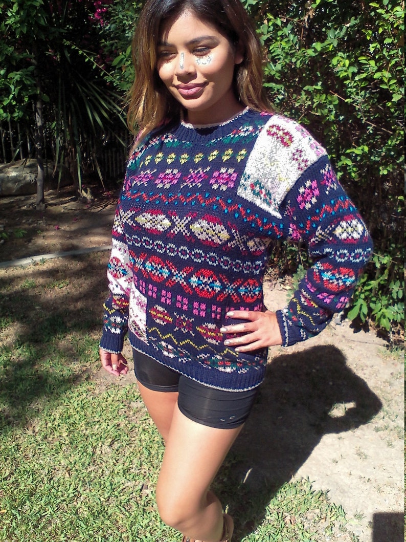 May include: A blue, white, and pink patterned sweater with long sleeves. The sweater has a fair isle design with geometric shapes and a white patch on the right shoulder.