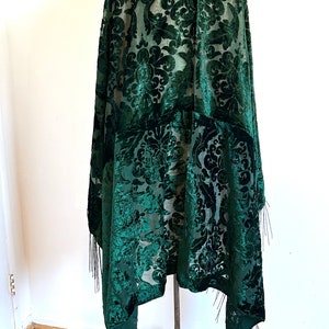 May include: A dark green velvet shawl featuring a sheer base with a raised, flocked damask pattern. The garment has a draped silhouette and is finished with long, thin fringe trim along the bottom edges, displayed on a metal stand.