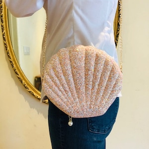 May include: A seashell-shaped crossbody bag in a shimmering peach color with a gold chain strap. The bag has a pearl charm and is worn over a white shirt and blue jeans. The bag's design resembles a scallop shell.
