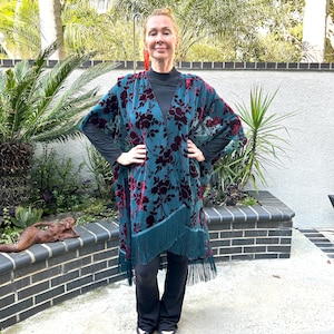 May include: A teal and burgundy floral patterned kimono with fringe trim. The kimono is worn over a black top and trousers. The image is taken outdoors with a brick wall and greenery in the background.