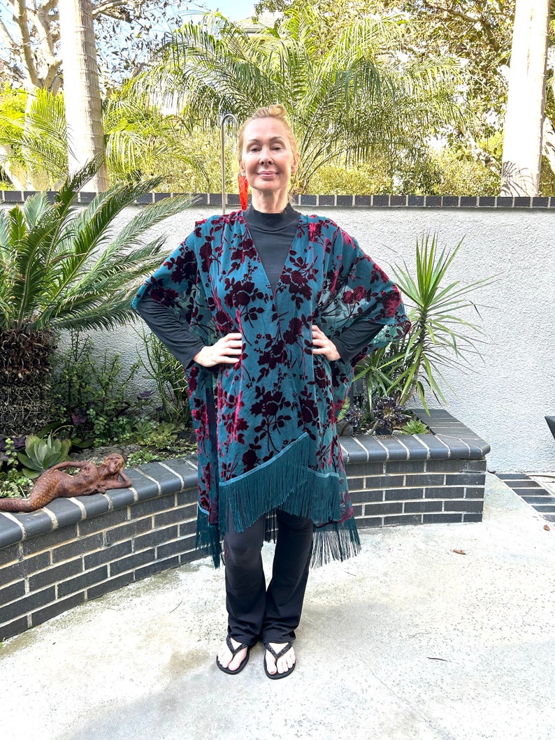 May include: A teal and burgundy floral patterned kimono with fringe trim. The kimono is worn over a black top and trousers. The image is taken outdoors with a brick wall and greenery in the background.