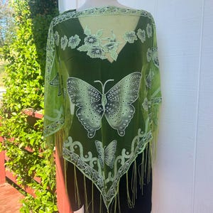 Puede incluir: Un chal de gasa verde con un diseño de mariposa y flores. El chal tiene un borde de flecos y está hecho de una tela ligera.