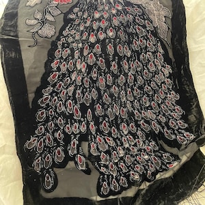 May include: Black velvet shawl with a peacock feather design. The shawl features a central peacock feather motif in black, gray, and red. The edges are trimmed with a black border and fringe. Floral accents in red and gray are also present.