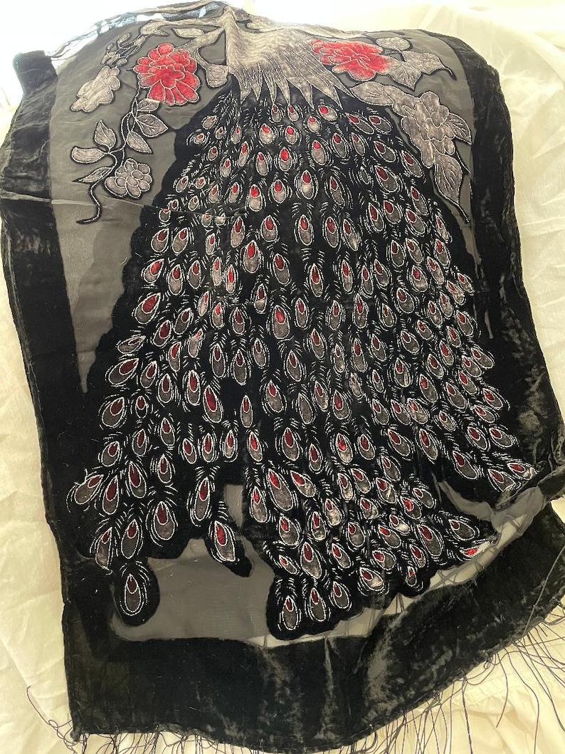 May include: Black velvet shawl with a peacock feather design. The shawl features a central peacock feather motif in black, gray, and red. The edges are trimmed with a black border and fringe. Floral accents in red and gray are also present.