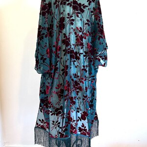 May include: A teal, sheer kimono-style robe with a V-neck and long sleeves. It has a floral pattern in a deep burgundy colour. The bottom hem is finished with a fringe. The robe is displayed on a metal stand.