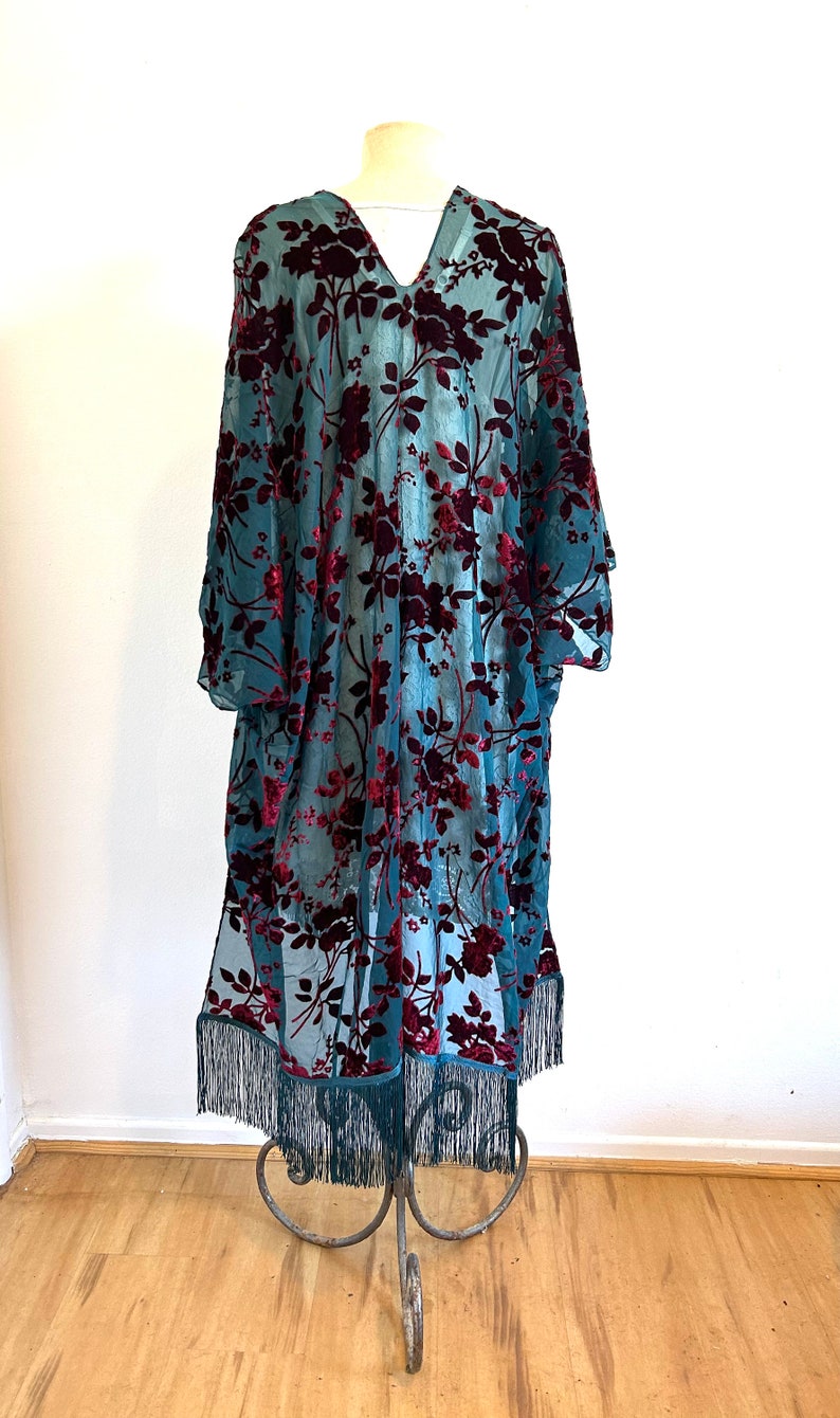 May include: A teal, sheer kimono-style robe with a V-neck and long sleeves. It has a floral pattern in a deep burgundy colour. The bottom hem is finished with a fringe. The robe is displayed on a metal stand.