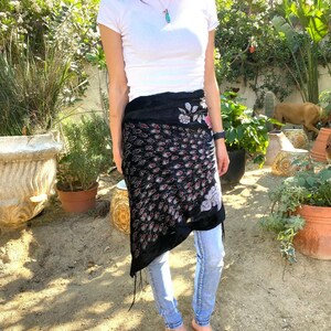 May include: A black wrap skirt with a pattern of red and white teardrop shapes and floral embroidery. The skirt is worn over light blue jeans. The skirt has a fringed edge.