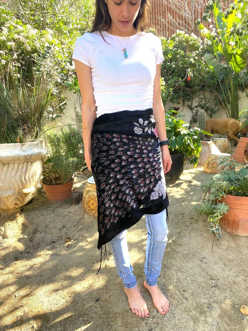 May include: A black wrap skirt with a pattern of red and white teardrop shapes and floral embroidery. The skirt is worn over light blue jeans. The skirt has a fringed edge.