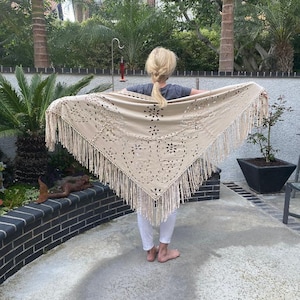 May include: A beige crocheted shawl with fringe trim and a cut-out design. The shawl is draped over a person's shoulders.