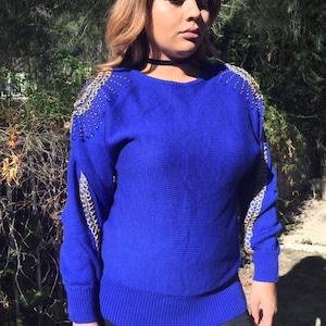 May include: A royal blue sweater with gold and blue beaded embellishments on the shoulders. The sweater has long sleeves and a round neckline.
