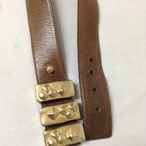May include: A brown leather belt with a gold metal buckle. The buckle is made up of three rectangular pieces with a raised design of circles and diamonds.