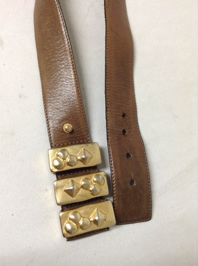 May include: A brown leather belt with a gold metal buckle. The buckle is made up of three rectangular pieces with a raised design of circles and diamonds.