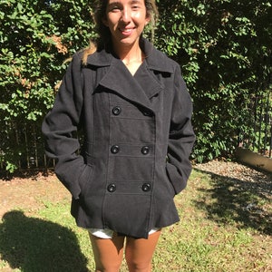 May include: A black double-breasted peacoat with a collar and pockets. The coat is worn by a person standing in front of a green bush.