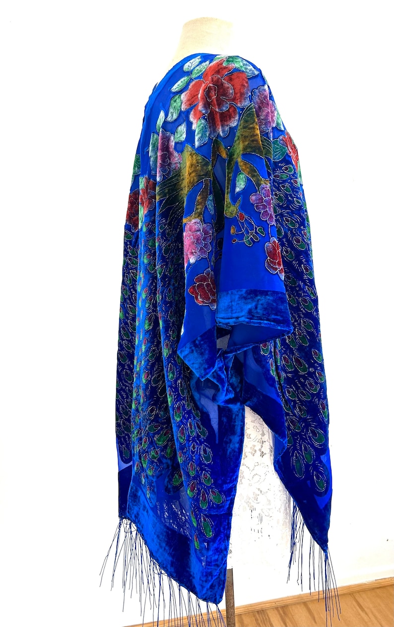 May include: A royal blue velvet shawl with a peacock feather design and floral embroidery. The shawl has a fringe trim and is draped over a white dress form.