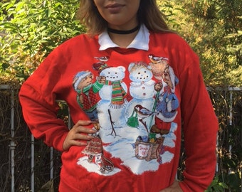 May include: Red Christmas sweater with a white collar and a snowman design featuring two bears and birds. The sweater has the text "Snow Day" on the bottom.