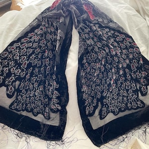 May include: A black scarf with a velvet border and fringe. The scarf features a pattern of stylized peacock feathers in shades of black, gray, and red. The scarf is draped on a white surface.