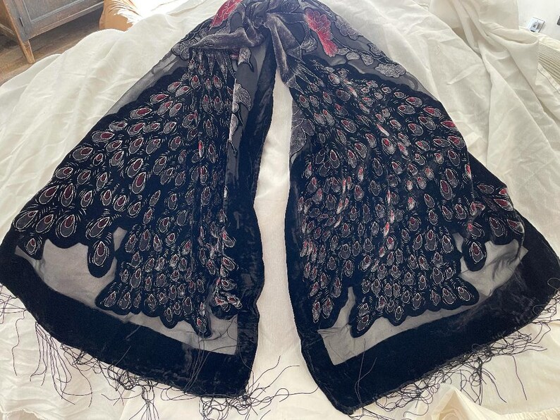 May include: A black scarf with a velvet border and fringe. The scarf features a pattern of stylized peacock feathers in shades of black, gray, and red. The scarf is draped on a white surface.