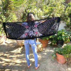 May include: A black sheer scarf with a peacock feather design in shades of black, pink, and white. The scarf is held up, showcasing the intricate details of the bird's feathers and floral accents. The scarf is rectangular and appears to be lightweight.