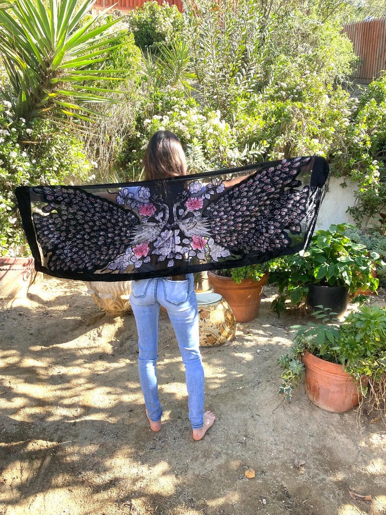 May include: A black sheer scarf with a peacock feather design in shades of black, pink, and white. The scarf is held up, showcasing the intricate details of the bird's feathers and floral accents. The scarf is rectangular and appears to be lightweight.