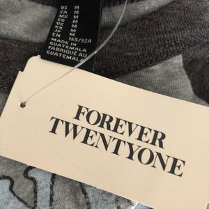 May include: Close-up of a gray Forever 21 t-shirt tag with the brand name and size information. The tag reads "FOREVER TWENTYONE" in bold black letters. The shirt has a light blue and gray pattern.