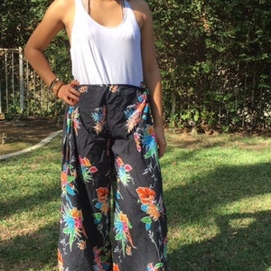 May include: A woman wearing a white tank top and black pants with a colorful floral print. The pants have a wide leg and are tied at the waist.