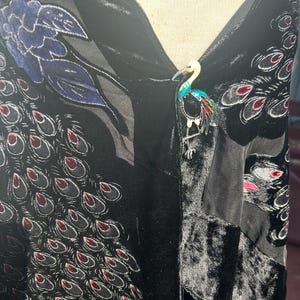 May include: A black velvet garment with a peacock feather pattern and a colorful bird brooch. The brooch features a white head, turquoise body, and red, yellow, and green accents. The garment has a floral design.