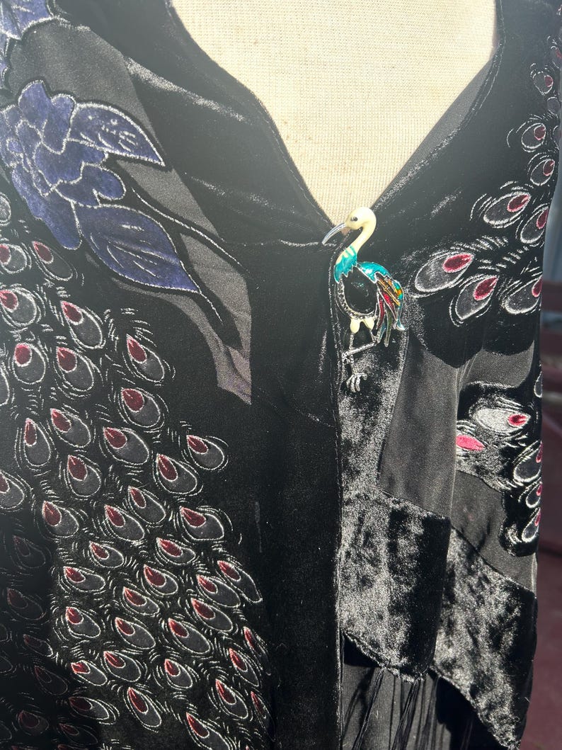 May include: A black velvet garment with a peacock feather pattern and a colorful bird brooch. The brooch features a white head, turquoise body, and red, yellow, and green accents. The garment has a floral design.