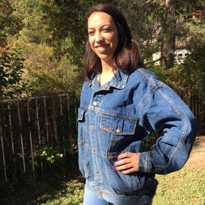 Pode incluir: Uma mulher vestindo uma jaqueta de jeans azul de lavagem clara. A jaqueta tem um fecho de bot&otilde;es na frente e mangas compridas. A mulher est&aacute; em p&eacute; em uma &aacute;rea gramada com &aacute;rvores no fundo.