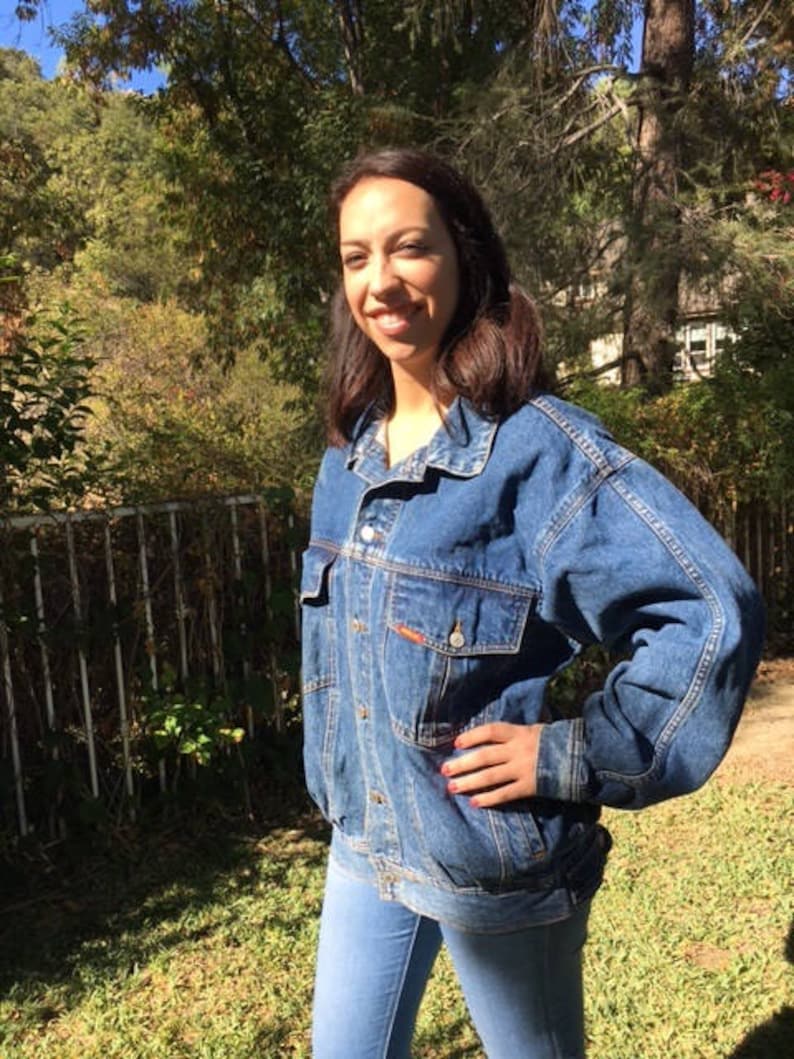 Pode incluir: Uma mulher vestindo uma jaqueta de jeans azul de lavagem clara. A jaqueta tem um fecho de bot&otilde;es na frente e mangas compridas. A mulher est&aacute; em p&eacute; em uma &aacute;rea gramada com &aacute;rvores no fundo.