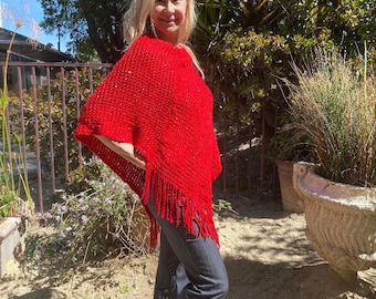 May include: A red knitted poncho with fringe detailing. The poncho is worn by a person with long blonde hair.