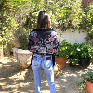May include: A black floral patterned top with pink and white flowers is worn with light blue jeans. The top has a sheer, delicate appearance. The jeans are fitted and have a classic five-pocket design. The person is standing in a garden setting.