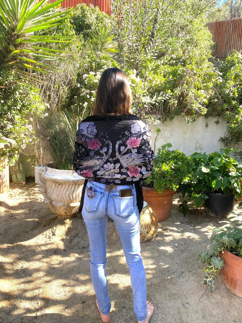 May include: A black floral patterned top with pink and white flowers is worn with light blue jeans. The top has a sheer, delicate appearance. The jeans are fitted and have a classic five-pocket design. The person is standing in a garden setting.