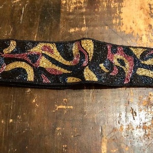 May include: A beaded belt with a black background and a swirling pattern in gold, pink, and blue.