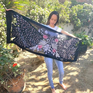 May include: A black shawl with a peacock design. The shawl features a detailed pattern of peacock feathers in shades of black, pink, and white. The design includes floral accents and a fringed edge. The shawl is held up, showcasing its full length.