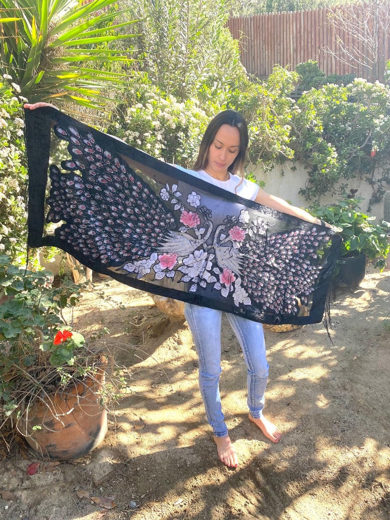 May include: A black shawl with a peacock design. The shawl features a detailed pattern of peacock feathers in shades of black, pink, and white. The design includes floral accents and a fringed edge. The shawl is held up, showcasing its full length.