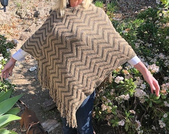 Op de afbeelding: Een vrouw draagt een bruine en beige chevron-gepatroneerde poncho met franjes, een bruine hoed, blauwe jeans en bruine schoenen. Ze staat in een tuin met een kleine bruine teckel.