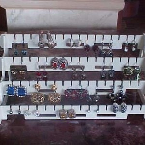 May include: White plastic earring organizer with four tiers. Each tier has slots for holding earrings. The organizer is filled with a variety of earrings, including silver, gold, and colorful stones.