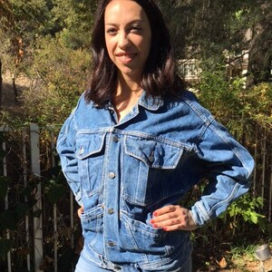 Pode incluir: Uma mulher vestindo uma jaqueta de jeans azul com bot&otilde;es e mangas compridas. A jaqueta tem um bolso grande na frente e um bolso com aba no lado esquerdo. A mulher est&aacute; vestindo jeans azul e tem as m&atilde;os na cintura.