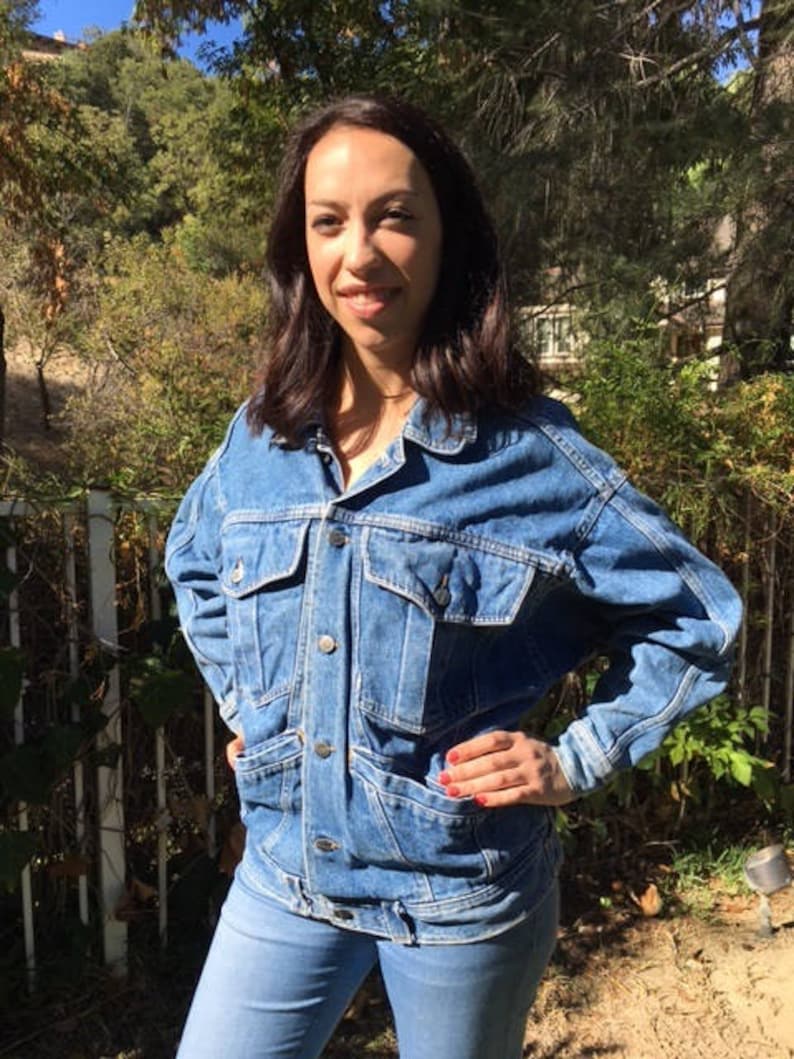 Pode incluir: Uma mulher vestindo uma jaqueta de jeans azul com bot&otilde;es e mangas compridas. A jaqueta tem um bolso grande na frente e um bolso com aba no lado esquerdo. A mulher est&aacute; vestindo jeans azul e tem as m&atilde;os na cintura.