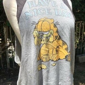 May include: Gray sleeveless t-shirt featuring Garfield the cat with the phrase "HAVE A NICE DAY" in light blue. The shirt has dark gray raglan sleeves and a distressed, vintage look.