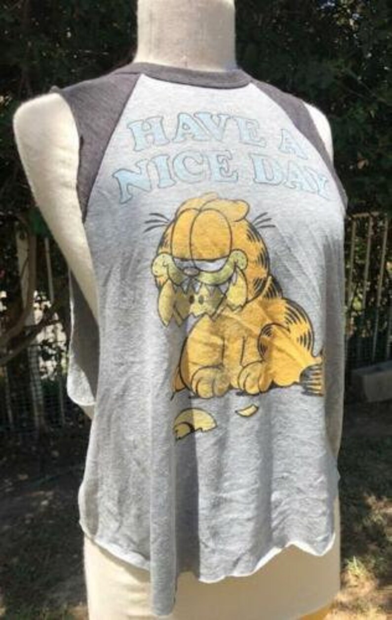 May include: Gray sleeveless t-shirt featuring Garfield the cat with the phrase "HAVE A NICE DAY" in light blue. The shirt has dark gray raglan sleeves and a distressed, vintage look.