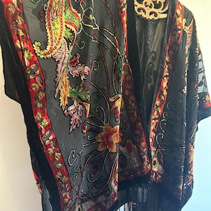 May include: A black kimono-style garment with a floral and paisley design. The kimono features a red, green, and yellow color palette, with a black fringe along the bottom. The garment is hanging on a wooden hanger.