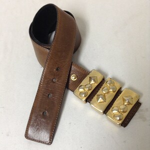 May include: A brown leather belt with a gold-toned buckle featuring multiple pyramid-shaped studs.